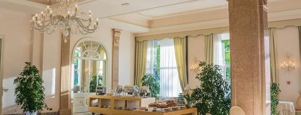 An elegant hotel dining room with a chandelier, tall windows and set tables